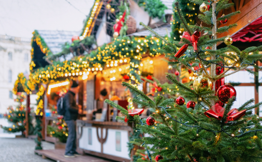 A decorated Christmas tree with red and gold ornaments and lights stands in the foreground of a blurry Christmas market scene