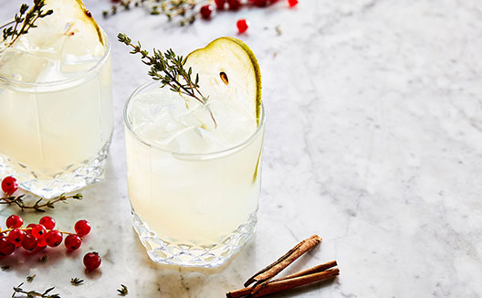 Two refreshing cocktails garnished with pear slices and thyme sprigs, with red currants and cinnamon on marble